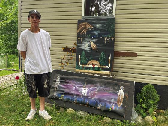 Artist Rik Yannott is pictured next to two of his paintings at his studio in Peshawbestown, both of which are inspired by nature and hisAnishinaabek heritage. The top painting, which features images of an eagle, the moon, and a traditional sweat lodge is on canvas, while the bottom painting with images of the Northern Lights and eagle feathers was completed on birch bark. Enterprise photo by Meakalia Previch-Liu